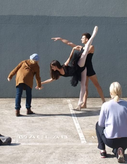 A dancer lifting one leg beside other performers in an outdoor paved space.