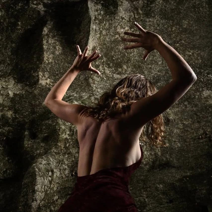 A dancer with raised arms beside a textured rock wall.