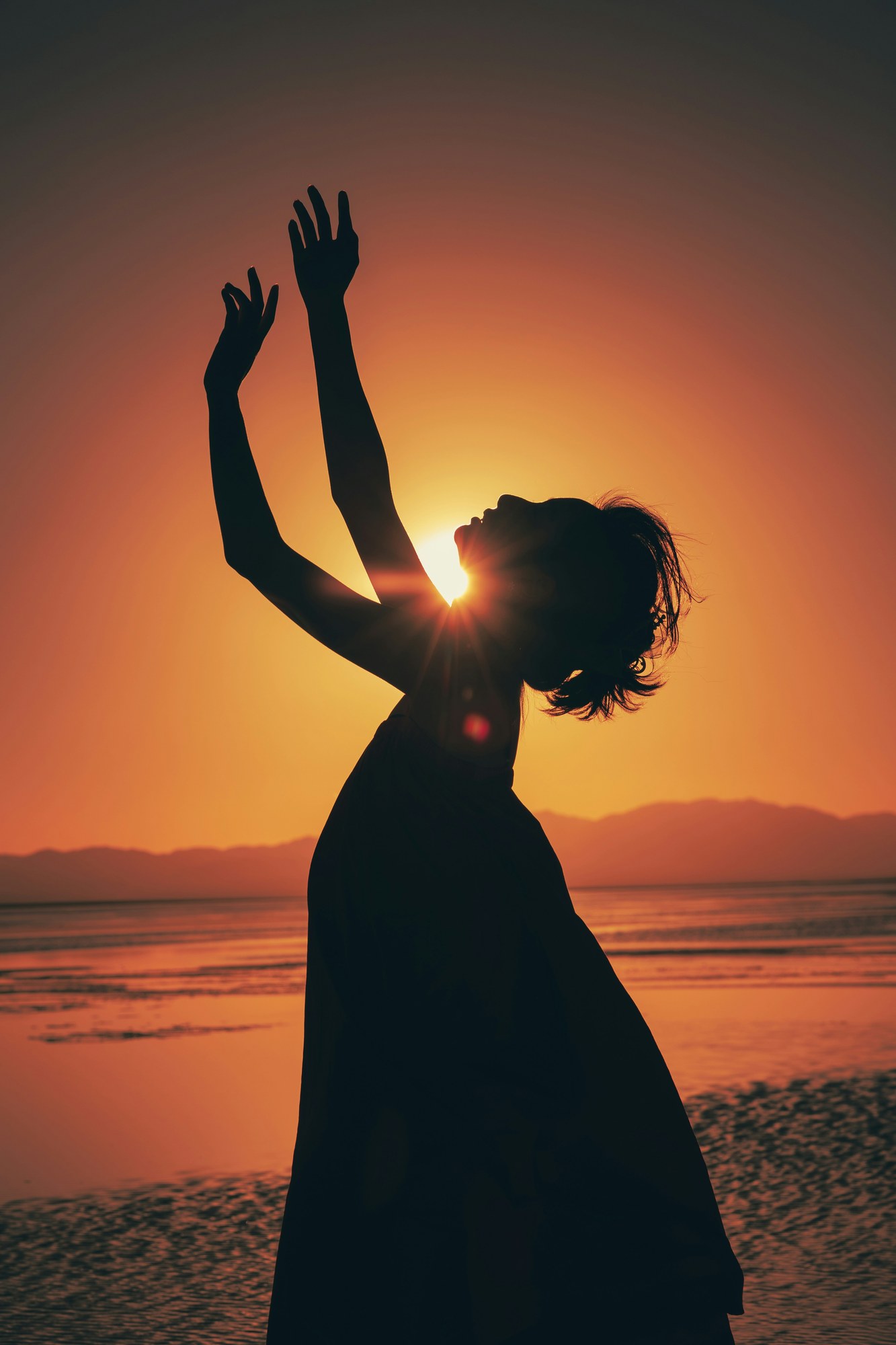 A woman raising her arms toward the sky beside the sea at sunset.